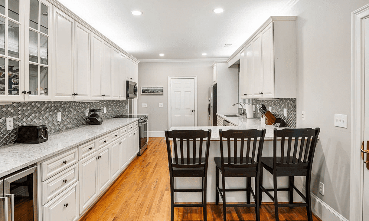 Modern kitchen with white cabinets, marble countertops, and a wooden floor. An island with three black chairs faces the stove and refrigerator. Mosaic tile backsplash decorates the walls.